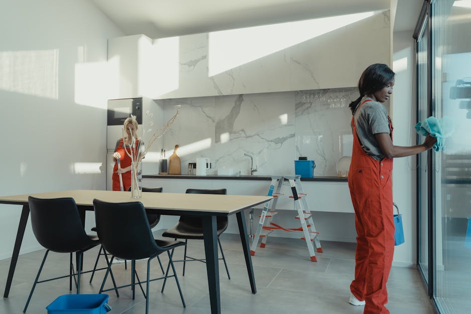A professional cleaner from Botany Bay Carpet Cleaners, dressed in protective coveralls, is engaged in surface cleaning inside a modern residential living room. The cleaner is using a long-handled squ