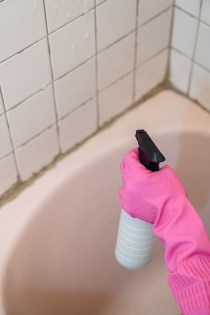A person wearing a bright pink rubber glove is holding a white spray bottle with a black nozzle, applying a cleaning solution to the edge of a pale pink bathtub. The bathroom features white tiled wall