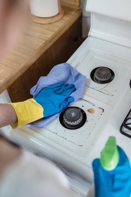 Close-up of a person wearing yellow and blue rubber gloves, using a light blue cleaning cloth to scrub the greasy, stained stovetop of a white oven in a domestic kitchen. The stovetop features two bla