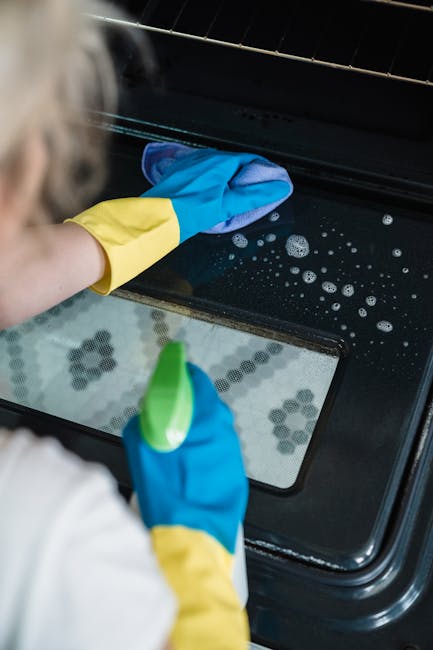 Close-up of a person wearing yellow and blue rubber gloves, using a light blue cleaning cloth to scrub the greasy, stained stovetop of a white oven in a domestic kitchen. The stovetop features two bla